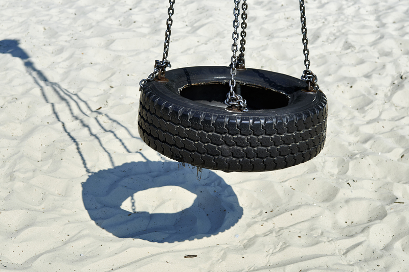 A black rubber tire swing with a textured tread pattern is suspended from metal chains attached to a circular frame, hanging above a sandy surface. The swing's shadow is cast onto the beige, fine-grained sand below, creating a clear circle of shadow. The swing is positioned centrally in the image, with the chains extending upward and out of the frame. The sandy area appears clean, with no other objects visible nearby. The scene suggests an outdoor play area or beach setting, with natural daylight illuminating the scene evenly. This image relates to rubbish and waste context in terms of the material (rubber) and possible removal or disposal processes by services such as Mattress Disposal in London, highlighting the importance of responsible waste management of outdoor play equipment or garden waste.