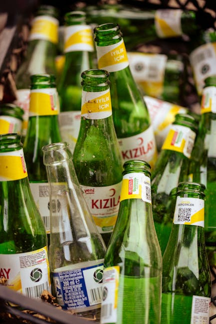 A close-up view of multiple green glass bottles with white and yellow labels, arranged in a scattered manner outdoors on a surface. The bottles are of similar size and shape, with some lying on their