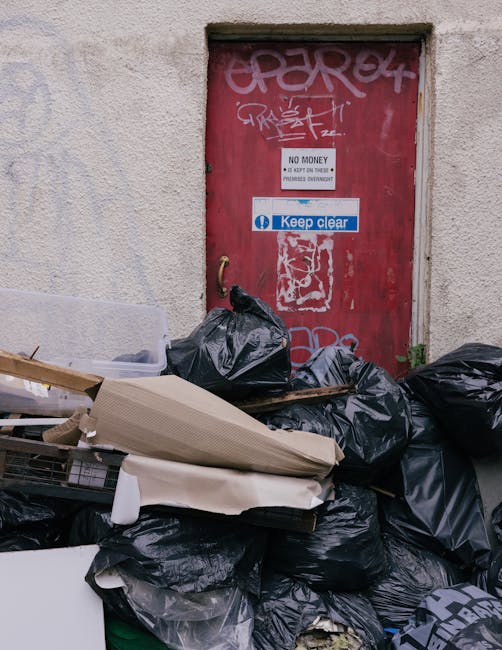 A collection of black garbage bags and discarded cardboard packaging piled in front of a red metal door on an external wall of a building. The door features various graffiti tags in white and pink pai