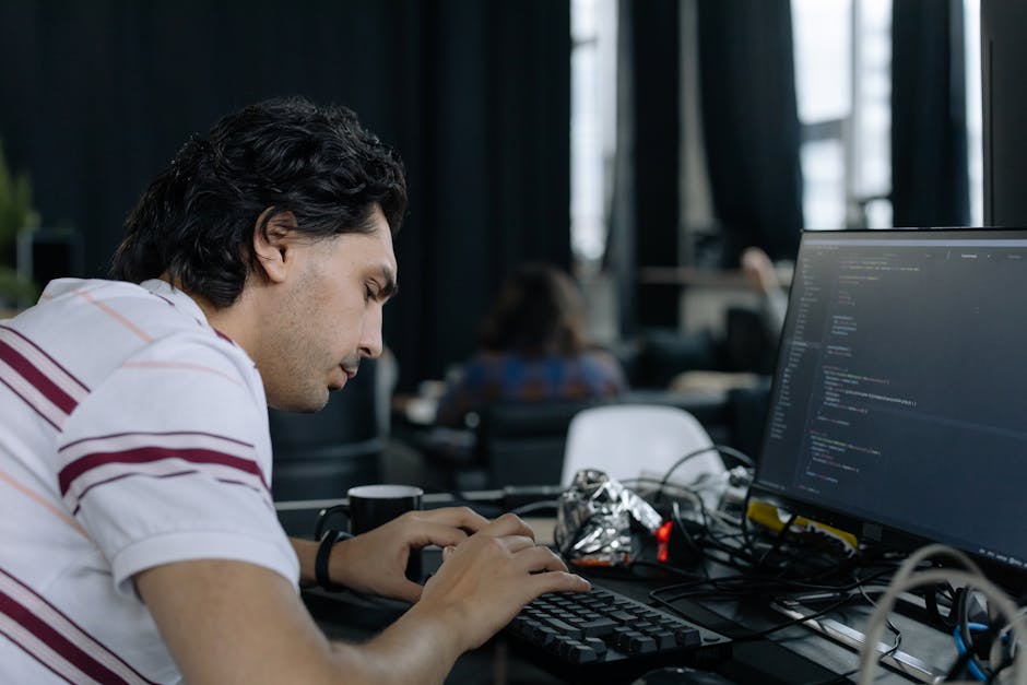 A young man with dark, wavy hair is seated at a cluttered desk in a modern office space, working intently on a computer with a large monitor displaying lines of code. He wears a white shirt with red a