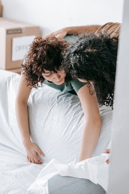 A minimalist interior bedroom scene featuring a neatly made white bed with a padded, upholstered headboard positioned against the right wall. The mattress appears unused, with a clean, smooth surface