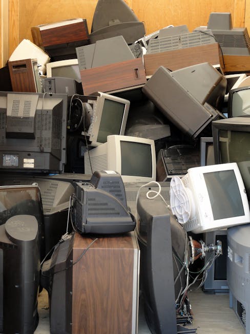 A large collection of discarded electronic devices, primarily comprising old computer monitors and CRT screens, stacked and piled together in an indoor environment. The monitors vary in size and desig